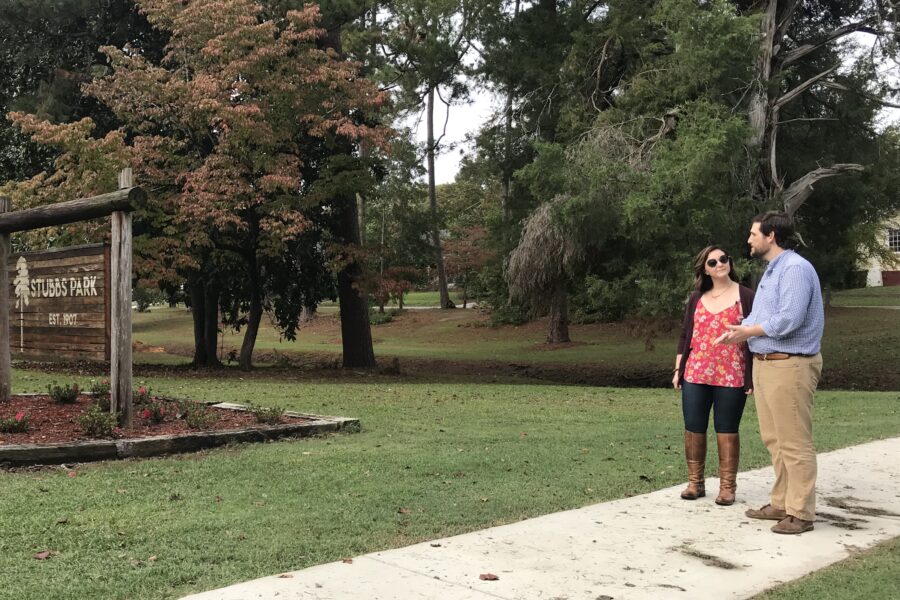 Two people walking along a sidewalk near the Stubbs Park entrance sign.