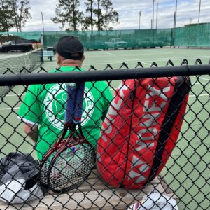 A man sits with his back to the camera and tennis equipment behind him and stares at the tennis court on Super Saturday