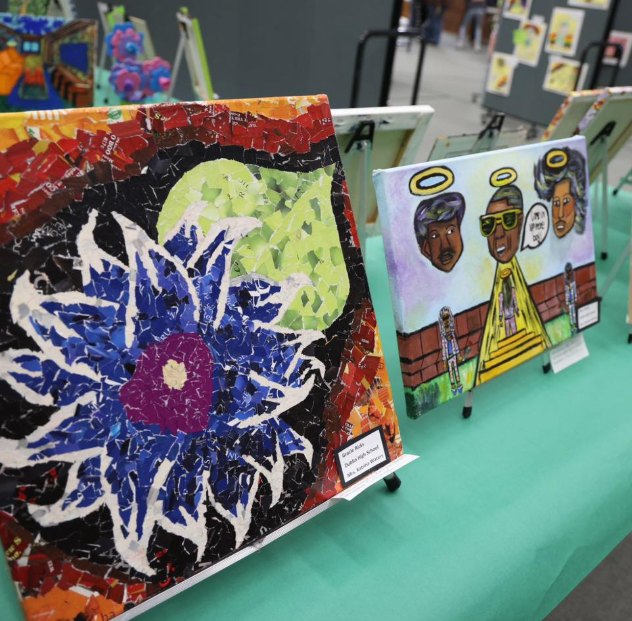 A painting of a spiky blue and purple flower with a large leaf and a painting of three faces with halos are displayed on a table at the St. Patrick's Student Art Show.