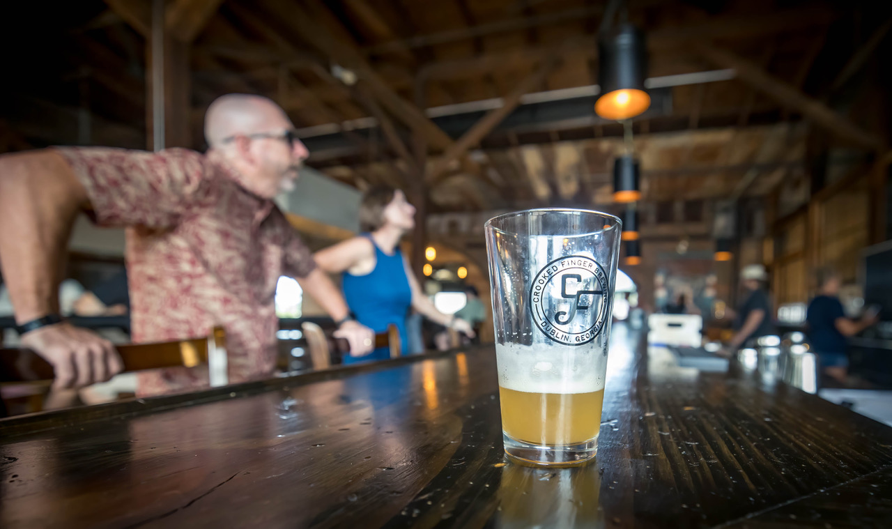A logo glass half-full of craft beer on a wood counter at Crooked Finger Brewing. Patrons wait to be served in the background.