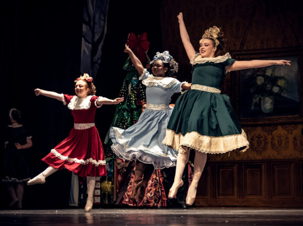 Three dancers dressed in vintage party dresses, with bows and ringlets in their hair, leap into the air in The Nutcracker