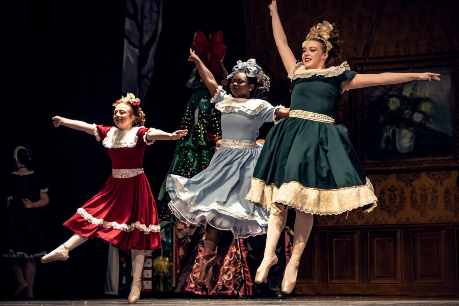 Three dancers dressed in vintage party dresses, with bows and ringlets in their hair, leap into the air in The Nutcracker