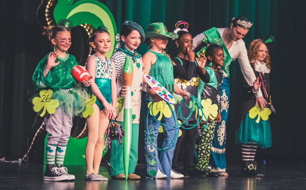 Little Girls Dressed as a wide Assortment of Leprechauns stand on a Stage in front of lit Shamrock Leprechaun Contest