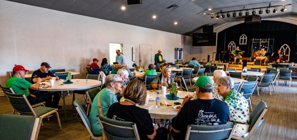 People decked out in green sitting at tables enjoying Corned beef & Cabbage Dinner while a band plays on a stage.