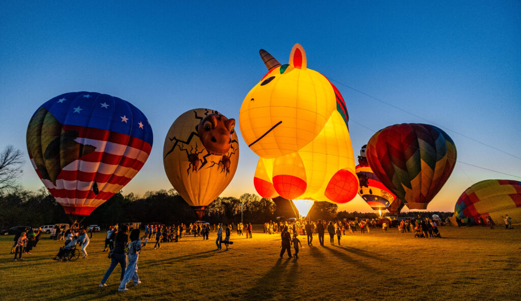 Multiple hot air balloons, including a unicorn and dinosaur egg, floating and glowing in Dublin, Georgia.