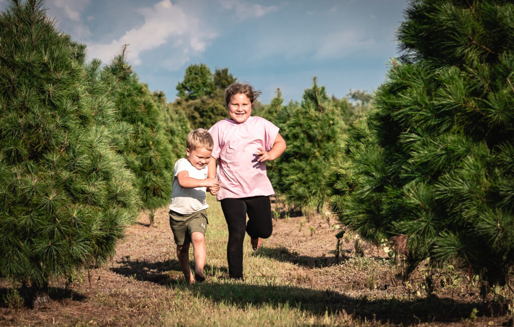 Two children running down a row of Christmas trees with hugs smiles on their faces.