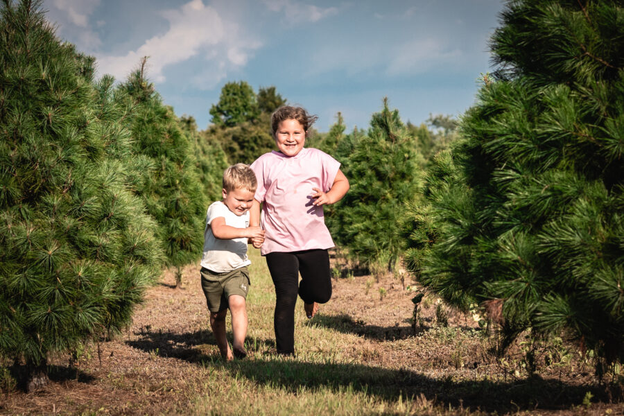 Two children running down a row of Christmas trees with hugs smiles on their faces.