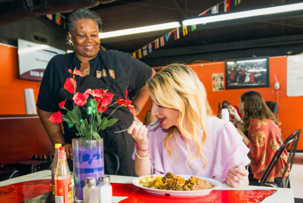 Community highlight Mama P standing at a table in her restaurant smiling at a patron enjoying her meal.