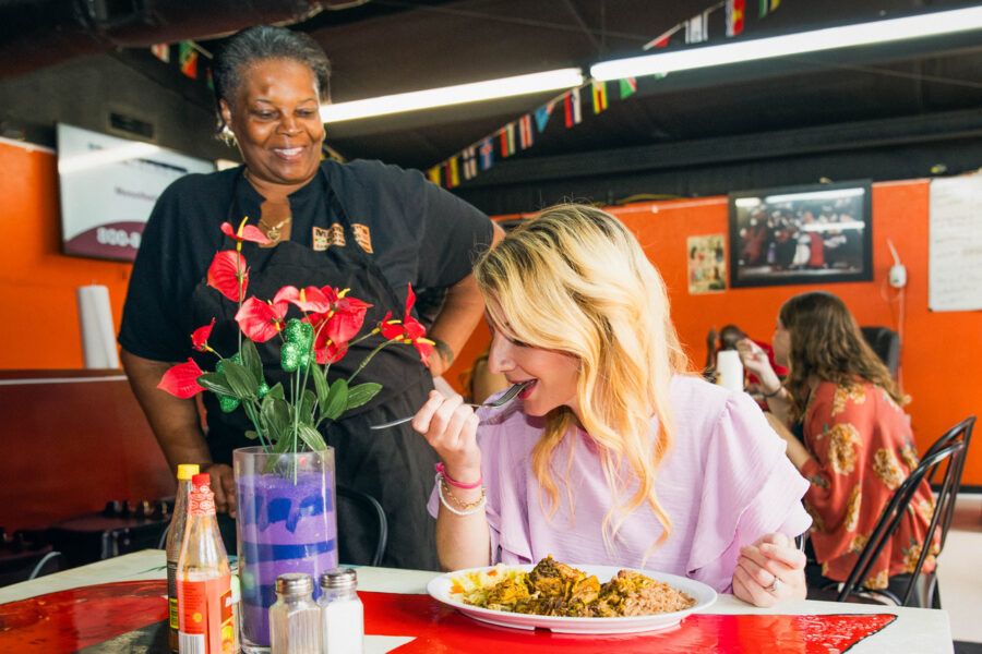 Community highlight Mama P standing at a table in her restaurant smiling at a patron enjoying her meal.