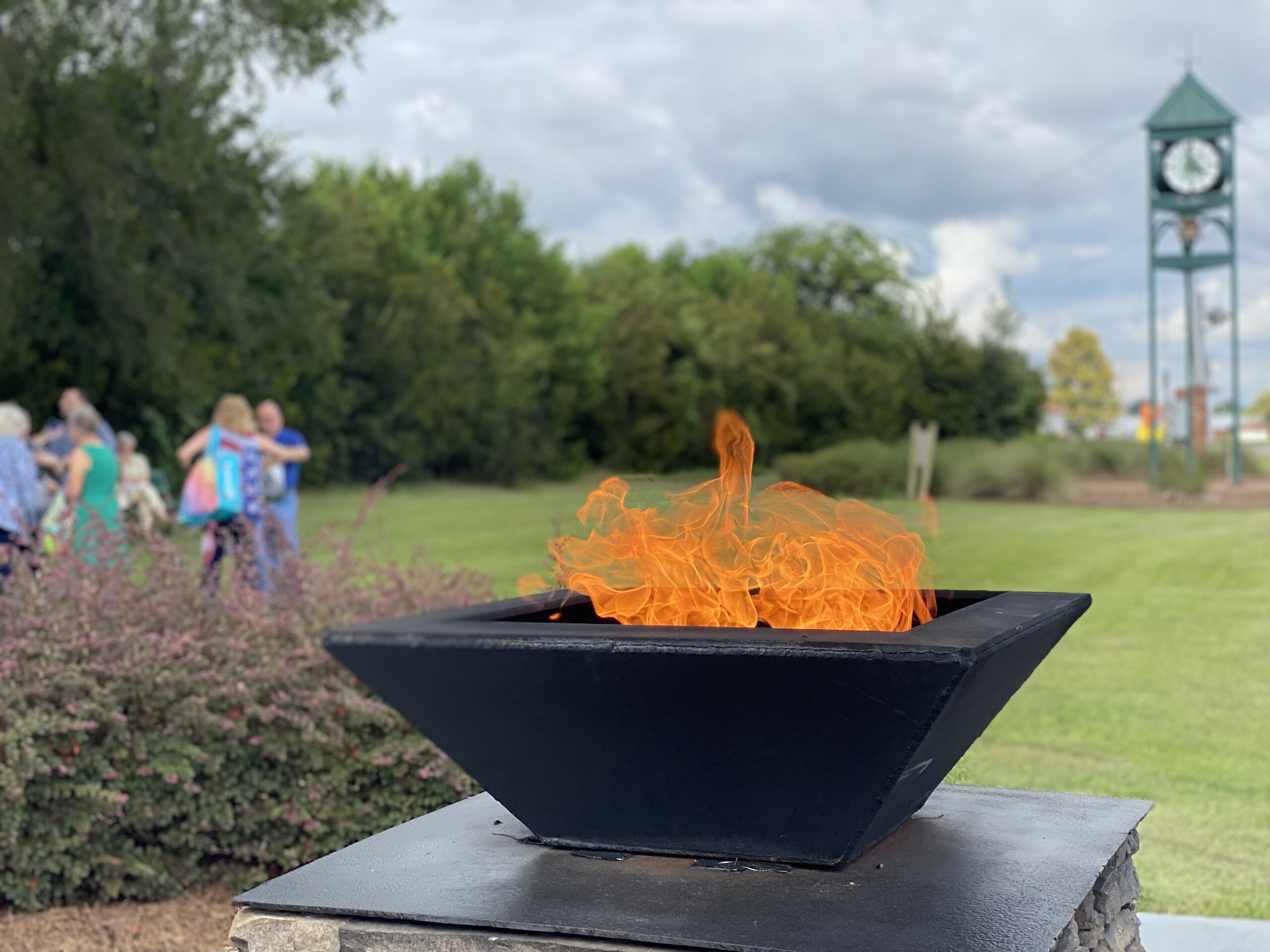 Everlasting flame in foreground and people attending Bells Of Freedom and Dublin's bell that is rung during the event in the background, all in Railroad Park.