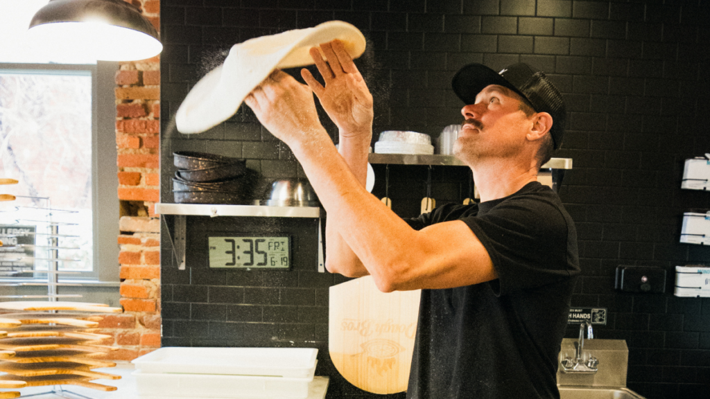 Owner of Dough Bros Brad Meeks spinning pizza dough above his head shaping a pizza crust.