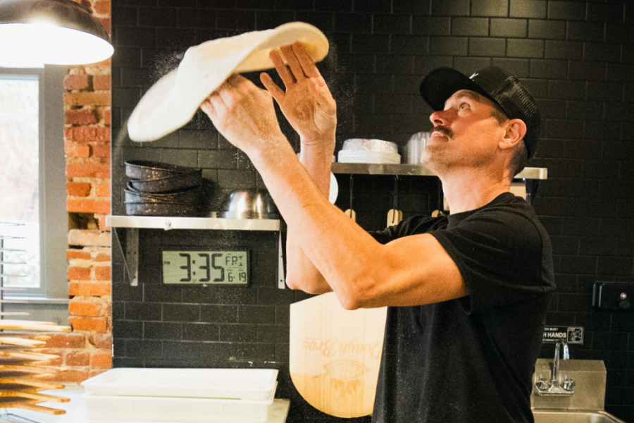 Owner of Dough Bros Brad Meeks spinning pizza dough above his head shaping a pizza crust.