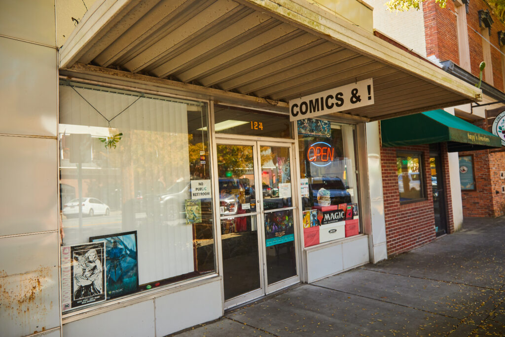 A storefront with large windows and an overhang with a metal sign reading Comics&!