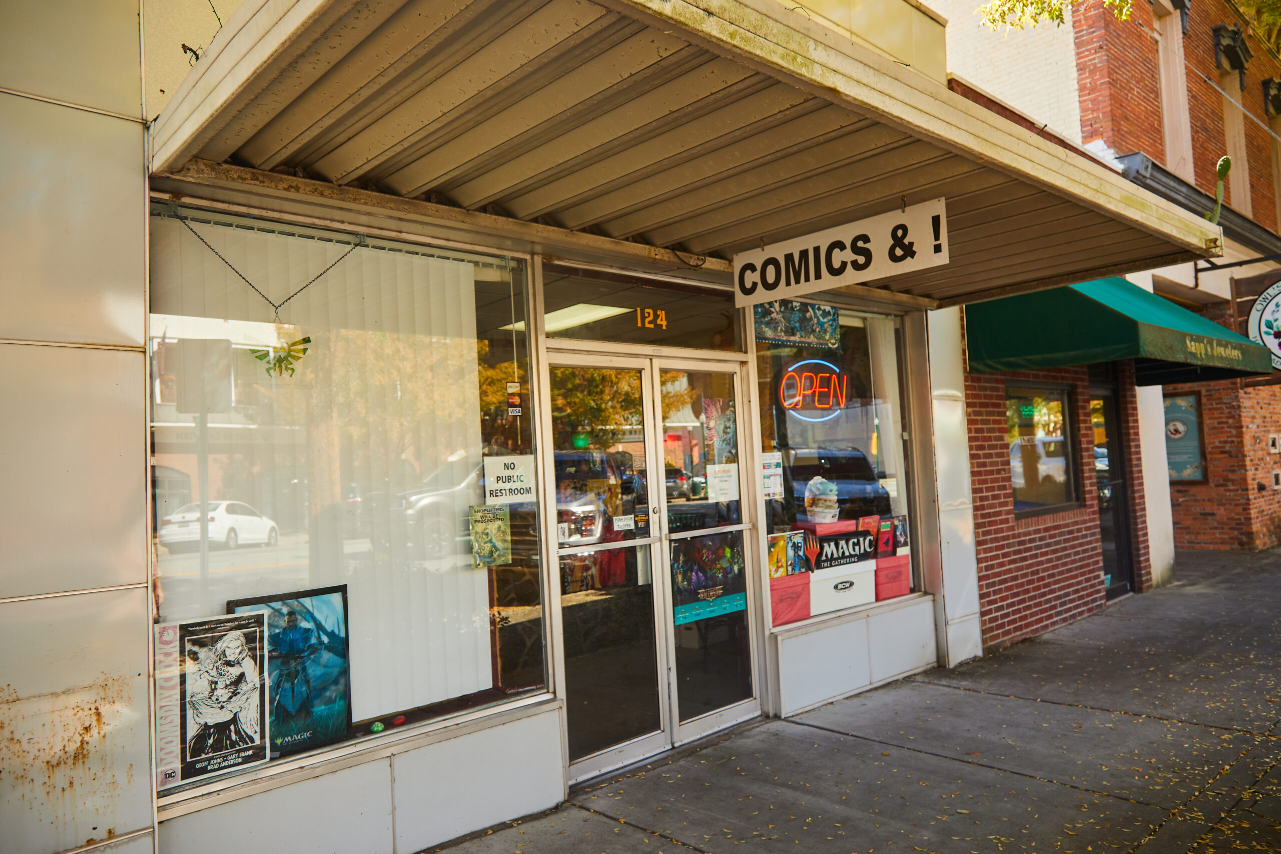 A storefront with large windows and an overhang with a metal sign reading Comics&!